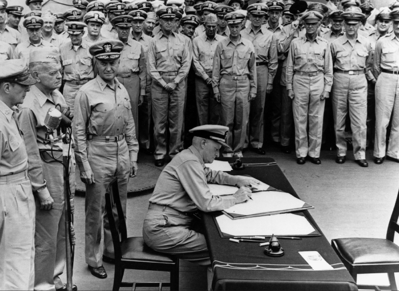 Fleet Admiral Chester W. Nimitz aboard USS Missouri in Tokyo Bay, signs Instrument of Surrender as United States representative, September2, 1945; Standing directly behind him are, left to right, General of the Army General Douglas MacArthur; Admiral William F. Halsey, U.S. Navy, andRear Admiral Forrest Sherman, U.S. Navy (U.S. Navy)
